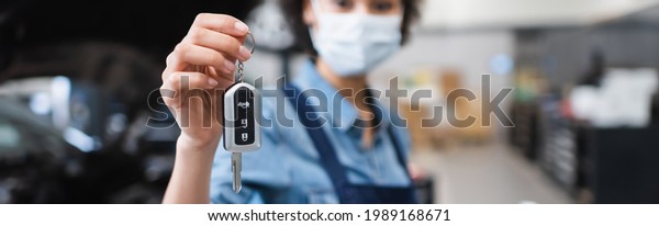 close up\
view of car key in hand of blurred young african american mechanic\
in protective mask in garage,\
banner