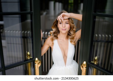 Close view of charming female model with stylish hairdo and makeup, wearing in vogue dress with flirty cutouts and V-neckline, standing opposite opened balcony door, posing and looking away