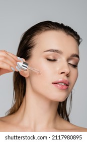 close up view of charming woman with closed eyes applying cosmetic serum isolated on grey - Shutterstock ID 2117980895
