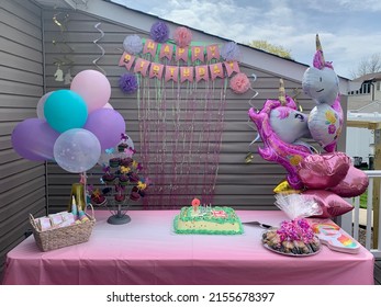 The close up view of a children's birthday party set up. The theme is unicorns with balloons, a banner, tissue paper flowers, cupcakes, cookies, plates, napkins, and an ice cream cake with candles.