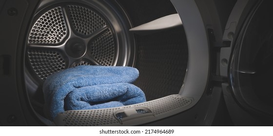 Close Up View Of A Clothes Dryer With Blue Towels Inside And An Open Door. Washing Machine In A Dark Room With A Brightly Lit Drum. Wide View