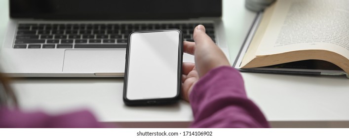 Close up view of college student using smartphone to relax while doing assignment with laptop on white table