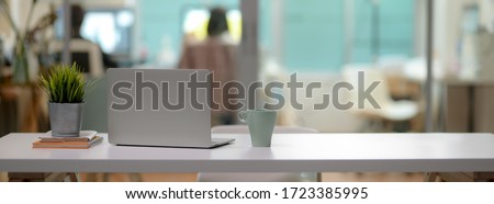 Close up view of comfortable office desk with laptop, mug, tree pot, office supplies and copy space on white table in glass partition office