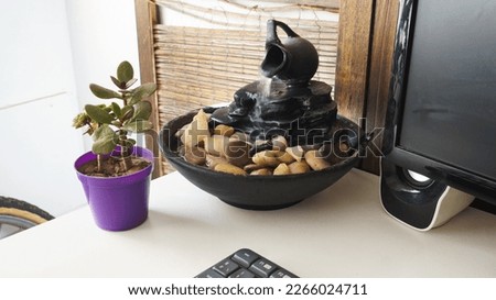   Close view of a decorative zen water fountain next to a keyboard, a monitor and succulent small plant. on top of a white wooden desk                             