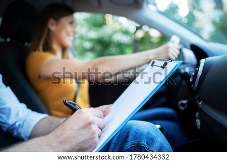 Close up view of driving instructor holding checklist while in background female student steering and driving car. Acquiring driver's license.
