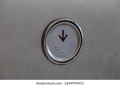 Close up view of an elevator button with downward arrow and braille dots on metal panel