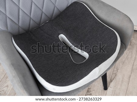 Close up view of ergonomic memory foam seat cushion, charcoal grey color with light grey trim, donut shape cutout for coccyx relief. Cushion rests on modern grey quilted chair, offering comfort for prolonged sitting.