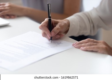 Close up view female hand holding pen signing legal paper seated at desk indoors, client receives services affirming document, hiring and human resources concept, put signature on employment contract