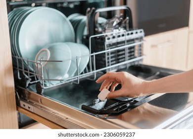 Close up view of female hand inserting dishwasher tablet into open built-in dishwasher machine with utensils inside in modern home kitchen. Household, housekeeping, domestic concept