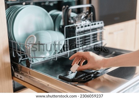 Close up view of female hand inserting dishwasher tablet into open built-in dishwasher machine with utensils inside in modern home kitchen. Household, housekeeping, domestic concept