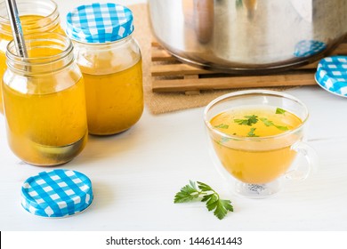 Close up view of a freshly poured glass of bone broth with herbs sprinkled on top surrounded by jars and a stock pot of freshly made bone broth.