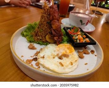 A Close Up View Of Fried Rice With Chicken Satay, Pickels, Vegetables And Fried Egg Served On A White Plate