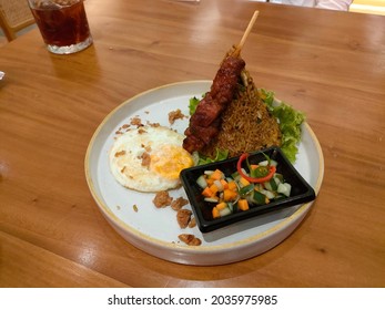 A Close Up View Of Fried Rice With Chicken Satay, Pickels, Vegetables And Fried Egg Served On A White Plate