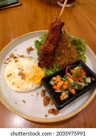 A Close Up View Of Fried Rice With Chicken Satay, Pickels, Vegetables And Fried Egg Served On A White Plate