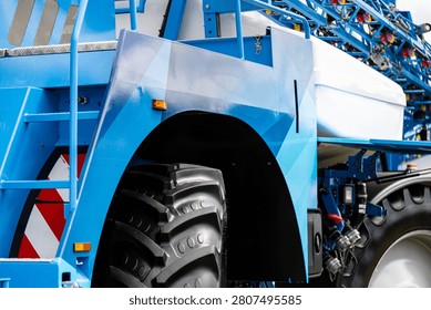 A close view of the front section of a self propelled crop sprayer shows a large deep tread wheel, blue structural panels, an access ladder, and parts of the spray boom with hoses and mounting elements. The machines robust frame and elevated design support field spraying operations in agriculture.