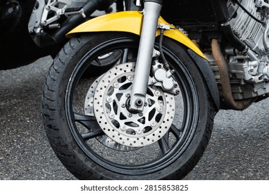 Close up view of front section of yellow and black sport motorcycle with black wheel, shiny brake disc, silver caliper, and suspension fork, parked on wet asphalt street