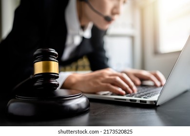 Close Up Of View Gavel With Female Lawyer Or Business Women Wearing Headset Using Laptop In The Background At A Law Firm, Selective Focus, Consult Legal Online, Justice And Law Concept.