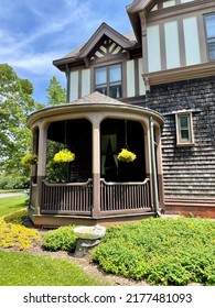 The close up view of a gazebo attached to a home. The small area is shaded, decorated with hanging plants, and has the view of a small bird bath.