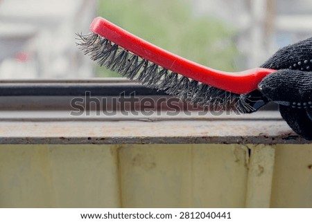 Close view of gloved hand using red wire brush to clean rust and old paint from metal balcony frame beside window, creating rough surface for fresh coating during outdoor maintenance