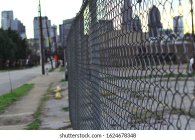 A close up view of a gray chain link fence that follows a sidewalk and street near a parking lot. The Dallas city skyline hangs in the background.