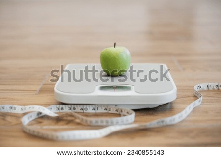 Close up view of green ripe apple being placed on bathroom scales while measuring tape lying on wooden floor nearby. Keeping active and eating balanced diet helping in maintaining healthy weight.