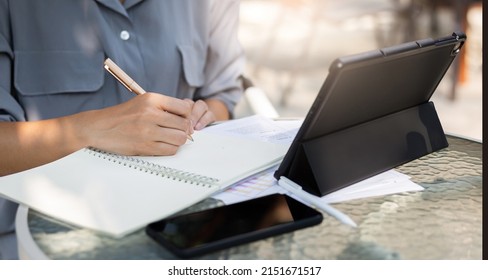 Close Up View, Hand Of Asian Woman Writing Her Plan On Notebook And Using Digital Tablet Working Online With Paperwork Of Investment On Glass Table