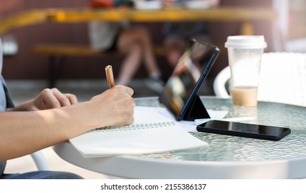 Close Up View, Hand Of Asian Woman Writing Her Plan On Notebook And Using Digital Tablet Working Online With Paperwork Of Investment On Glass Table