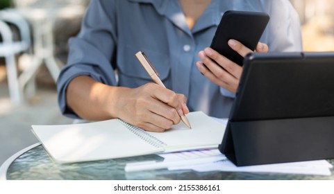 Close Up View, Hand Of Asian Woman Writing Her Plan On Notebook And Using Mobile Phone With Tablet Computer Working Online With Paperwork Of Investment On Glass Table