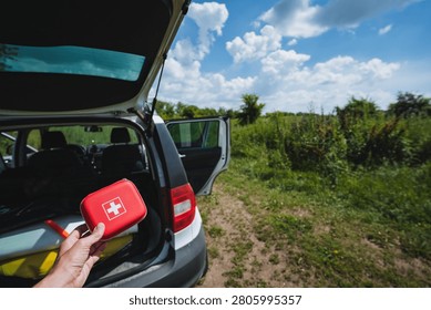 Close view of hand holding compact red first aid kit with white cross beside open hatchback trunk of white car parked on grassy dirt path near green field under bright blue sky with scattered clouds