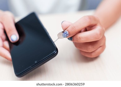 Close up view of a hand using a SIM card ejector tool on a smartphone on a light wooden table.