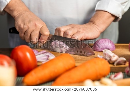 Close up view of the hands of an adult woman cutting onion with a kitchen knife on a wooden board for lasagna preparation.
