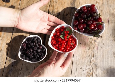 Close view of hands placing three heart shaped bowls filled with ripe cherries, glossy redcurrants and plump blackberries on weathered wooden table, with soft morning sunlight and gentle shadows