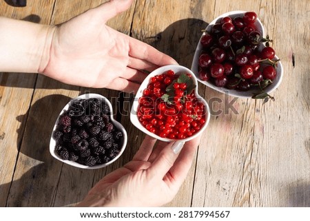 Close view of hands placing three heart shaped bowls filled with ripe cherries, glossy redcurrants and plump blackberries on weathered wooden table, with soft morning sunlight and gentle shadows