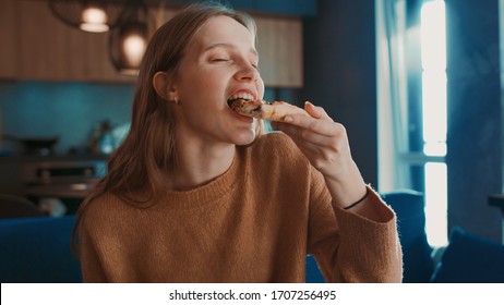 Close up view of happy hungry pretty blondehair girl sitting on blue sofa at home and eating tasty pizza, enjoying and smiling.
