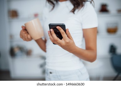 Close up view. Holds cup of drink in hand. Woman in white clothes with her smartphone resting in living room.