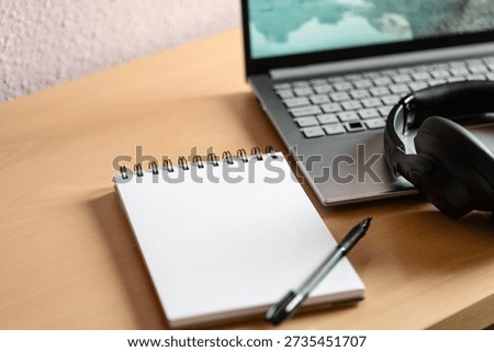 Close view of a home study desk representing lifelong learning and online education. Notebook, blank paper with copy space, pen, laptop keyboard and headphones on a minimal workspace photographed at a 45 degree angle.