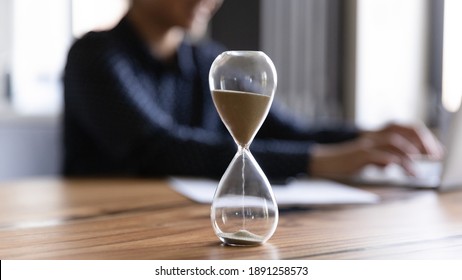 Close up view of hourglass stand on wooden home office measuring time, woman busy using computer. Female employee on background work on laptop, try to meet deadline. Efficiency concept. - Shutterstock ID 1891258573