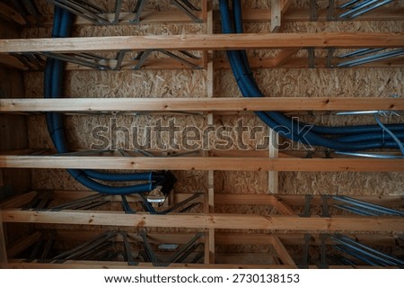 Close view inside shows OSB sheathing, solid joists, metal web trusses carrying blue corrugated ventilation ducts and conduit toward a distribution box during daylight rough in.