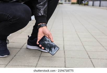 Close Up View Of Kneeling Man In Jeans And Shoes Picking Up Broken Phone On Paved Sidewalk Outdoors