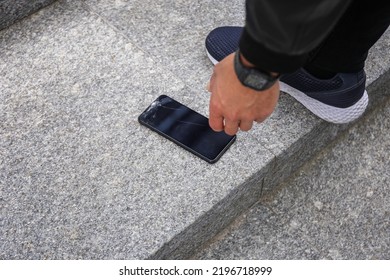 Close Up View Of Kneeling Man In Jeans And Shoes Picking Up Broken Phone On Paved Sidewalk Outdoors