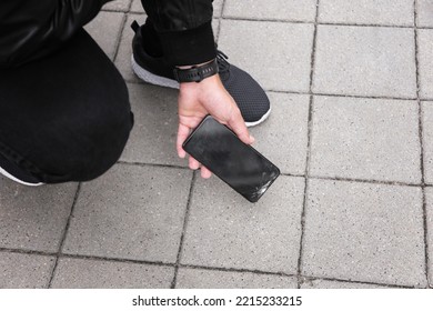 Close Up View Of Kneeling Man In Jeans And Shoes Picking Up Broken Phone On Paved Sidewalk Outdoors