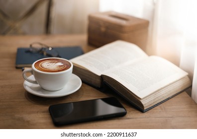 Close up view, Latte coffee in white cup with old book and smart phone on wooden table near bright window. blurred background, eyeglasses on blue note book and pen, vintage color tone - Powered by Shutterstock