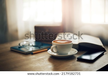Close up view, Latte coffee in white cup and smart phone on wooden table near bright window. blurred background with book, eyeglasses on blue note book and pen, vintage color tone