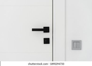Close up view of light switch on the white wall near modern minimalist door with black handle. Contemporary interior design details at home, apartment or office