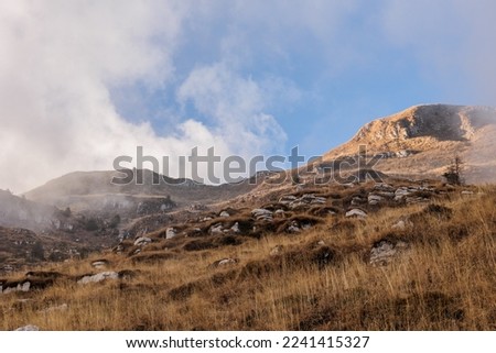 close up view, looking from the bottom to the top, of a mountainside covered with dry and gold-colored grass, full of rocks and stones. clouds passing above the mountain ridge and revealing the sky