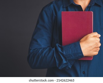 Close View Of Man In Blue Shirt Holding A Book With An Empty Red Cover In Hand While Standing On A Gray Background. Free Space For Your Mockup Of The Reading Book Concept