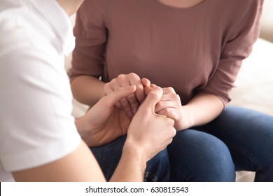 Close up view of man holding hands of a woman sitting on sofa indoors, love confession or marriage proposal, expressing support and comforting, asking for forgiveness and apologizing