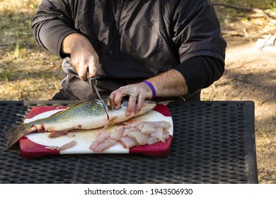 A close up view of a man sitting at a table outdoors cutting a fish into small chunks on a cutting board.