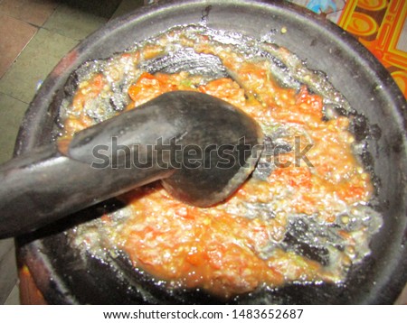 Close up view of Mengulek ulek sambal sambel or crush grind grill red hot small big chili, terasi, tomat tomato on cobek or stone plate mortar ulekan pestle oelek. Eat with dish meal spicy red color
