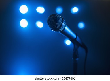 A close up view of a microphone and stand.  The background is blue and has several spotlights. Horizontally framed shot.